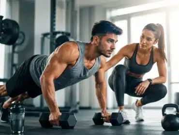 Fit male athlete doing dumbbell push-ups while female personal trainer coaches him in a modern fitness gym with strength training equipment.